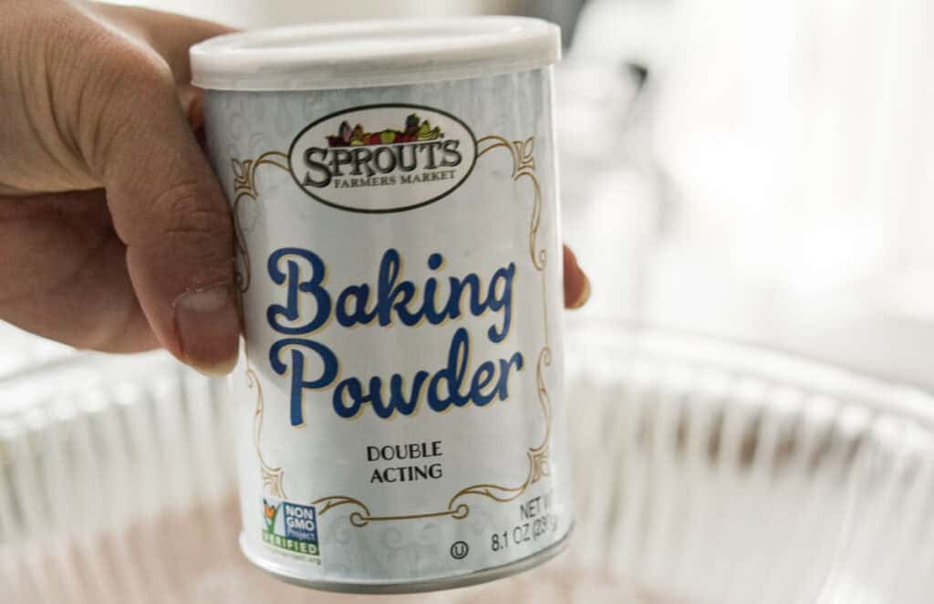 A hand holds a can of Sprouts Farmers Market Baking Powder over a metal baking pan. The label indicates it is double acting and non-GMO verified. The can has a white lid and decorative design.